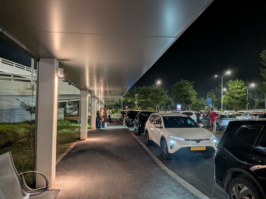 夜のノイバイ空港P1駐車場のGrab乗り場。屋根つきの歩道でスーツケースを持って待つ人たちと、車道に縦列で停まる黄色ナンバーの車