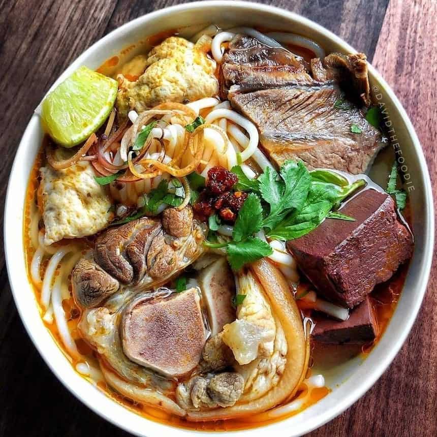 ベトナム中部フエの伝統料理「ブンボーフエ(Bún bò Huế)」。豚足、牛肉の薄切り、血のゼリー、カニカマがのった、オレンジ色のスープが特徴的な、パクチーやレモン、サテ(唐辛子ソース)を添えた食欲をそそる一品。