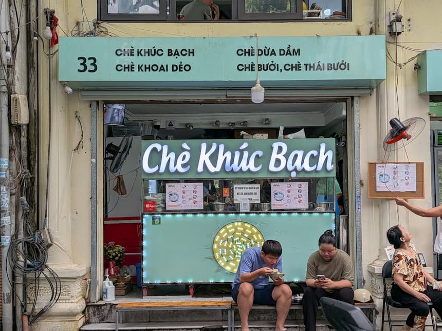 ハノイのクックバック専門店「Chè Nhà Suvy」（33 Quang Trung）の店頭。ミントグリーンの看板にチェー・クックバックやチェー・ズアザムのメニューが並び、店先の椅子で地元客がチェーを楽しんでいる。