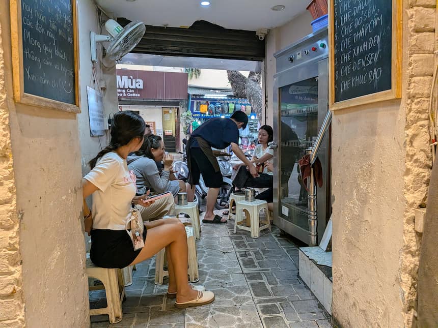 ハノイの路地裏にある小さなチェー(ベトナム風ぜんざい)店。プラスチックの椅子に座り、黒板に書かれた伝統的なチェーのメニューを楽しむ地元の人々の様子。
