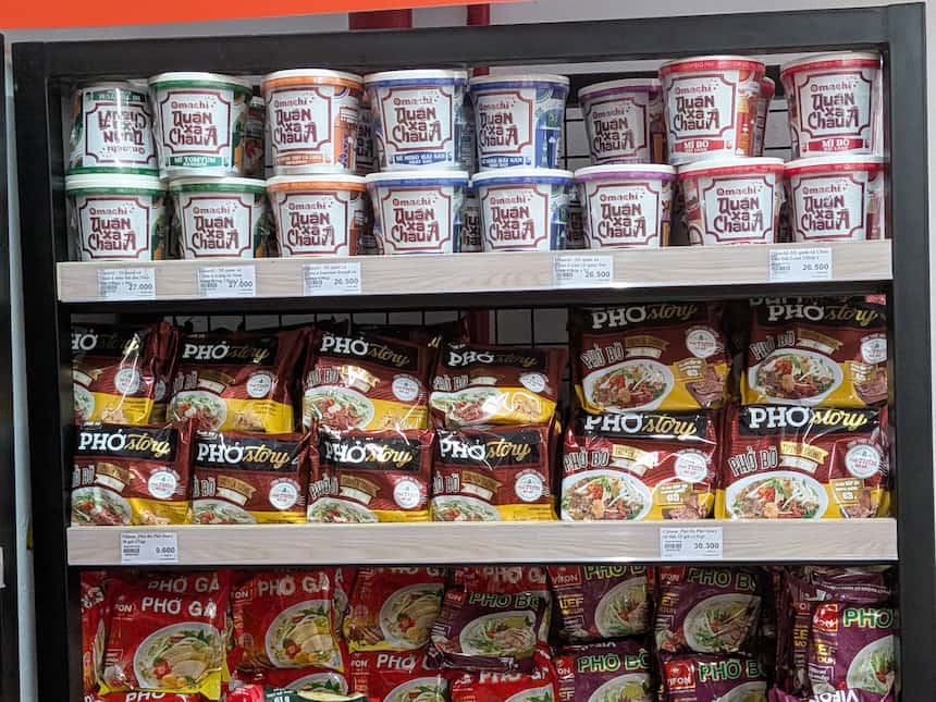スーパーマーケットの棚に並ぶインスタントラーメン（Omachi）とインスタントフォー（Pho Story）。種類豊富なカップ麺や袋麺が整然と陳列されています。