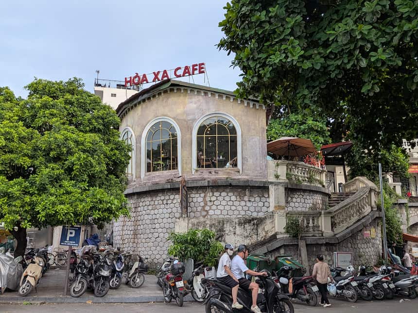 ロンビエン駅構内にあるHoa Xa Cafe(ホアサーカフェ)の外観。アーチ型の窓が並ぶ円形の旧駅舎の上部に赤い「HOA XA CAFE」の看板、手前にバイクが並ぶ。