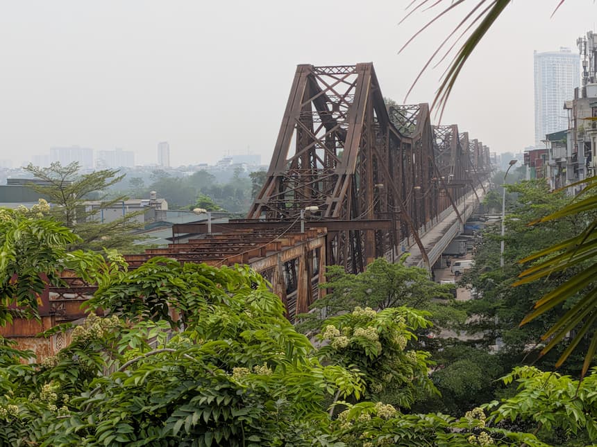 ハノイの歴史的建造物であるロンビエン橋（Long Biên Bridge）。赤茶色の鉄橋が紅河に架かっており、周囲の緑豊かな風景とともにその姿を捉えています。