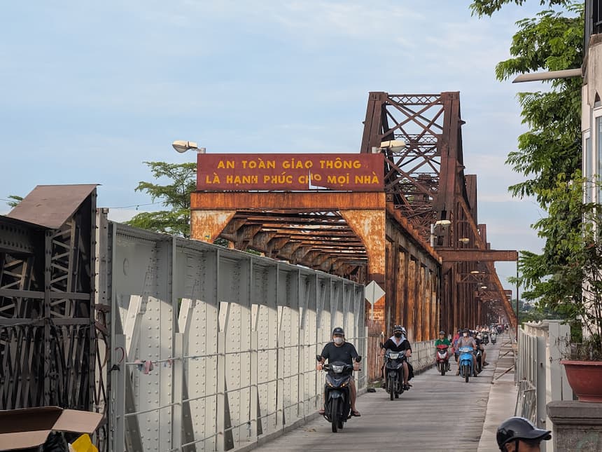 2026年5月に再開通したロンビエン橋。新しく舗装された路面をバイクが走り、手前に「AN TOÀN GIAO THÔNG(交通安全)」の赤い標識と錆びた鉄骨トラスが見える。