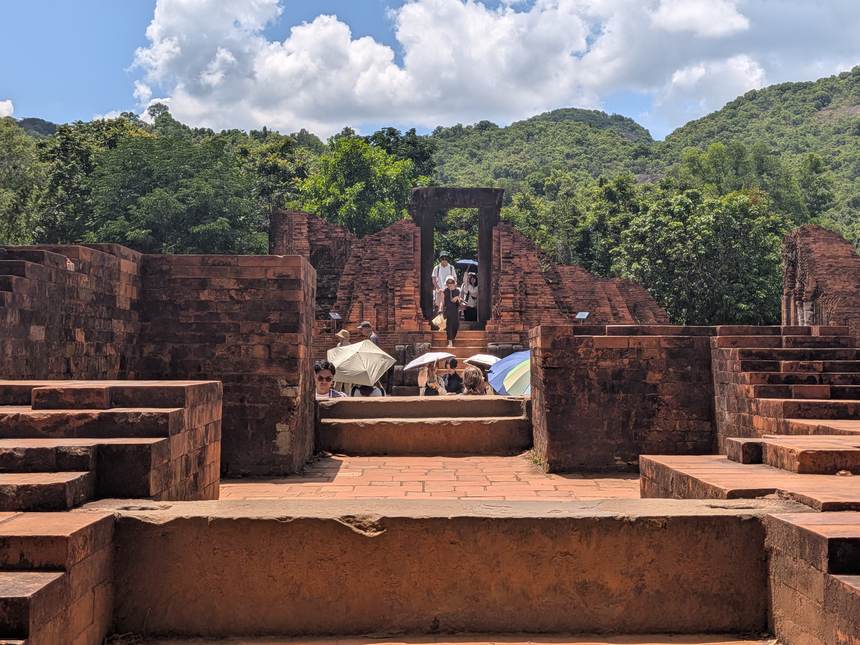 中部ベトナムの世界遺産・ミーソン遺跡。青空の下、日傘をさした観光客が炎天下の赤レンガ遺跡を見学している。中部の乾季は日差しが強い。