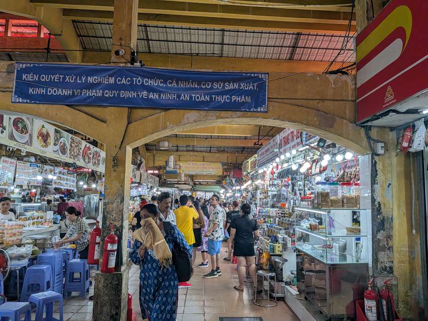 ベトナムの屋内市場の通路。両脇に食料品や雑貨の露店が並び、買い物客で賑わう。