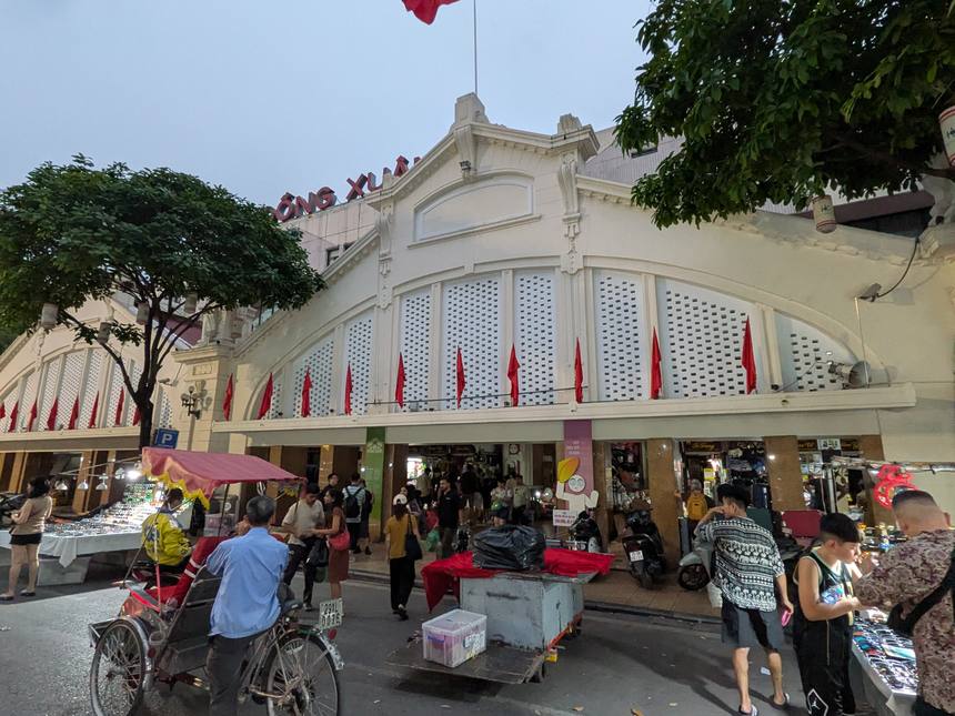 ハノイの旧市街にある大型市場の正面外観。コロニアル様式の建物にベトナム国旗が掲げられ、前をシクロや露店、買い物客が行き交う。