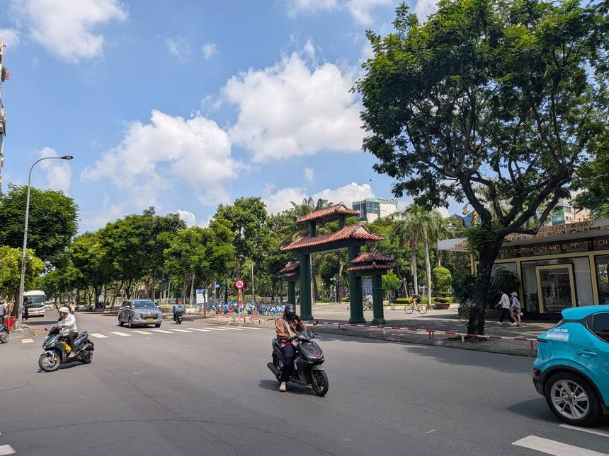 晴れた乾季のホーチミン市内。青空の下、公園の門と並木道のある大通りをバイクが行き交う