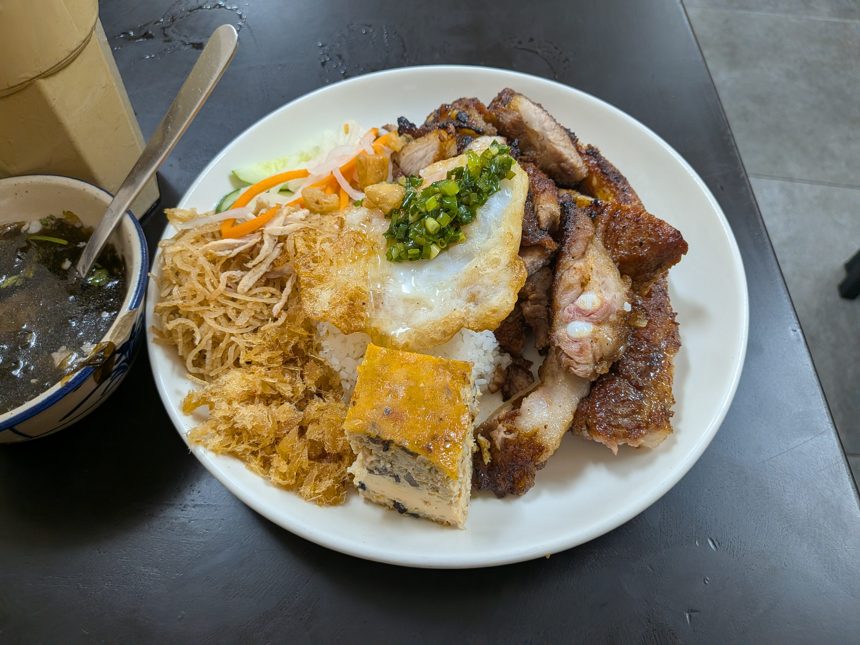 ベトナムの定番料理「コムタム(broken rice)」の一皿。白米の上にグリルポーク、揚げ卵、蒸し卵ケーキ、千切り豚皮などが盛り付けられ。