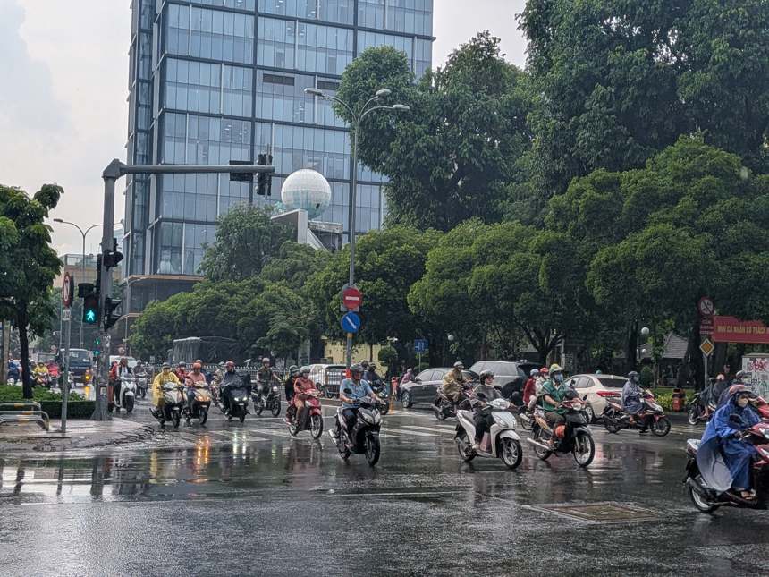 雨上がりで路面が濡れたホーチミン市の交差点を、無数のバイクが行き交う。レインコート姿のライダーも見える、活気あるベトナムの街の風景。