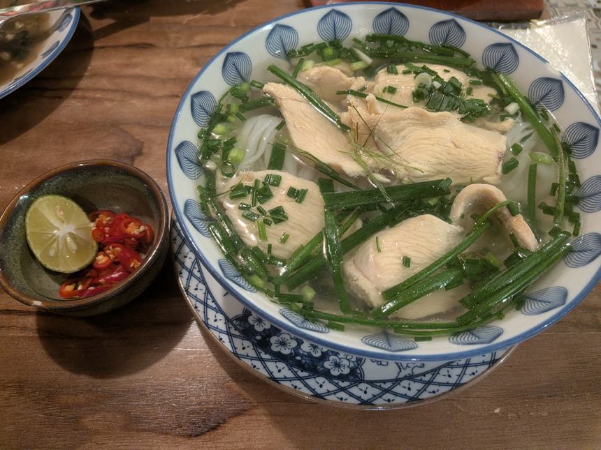 青と白の花柄の器に盛られたフォーガー(鶏肉のフォー)のベトナム料理。透き通ったスープに米麺、鶏肉、ニラが入り、小皿にはライムと唐辛子が添えられている。