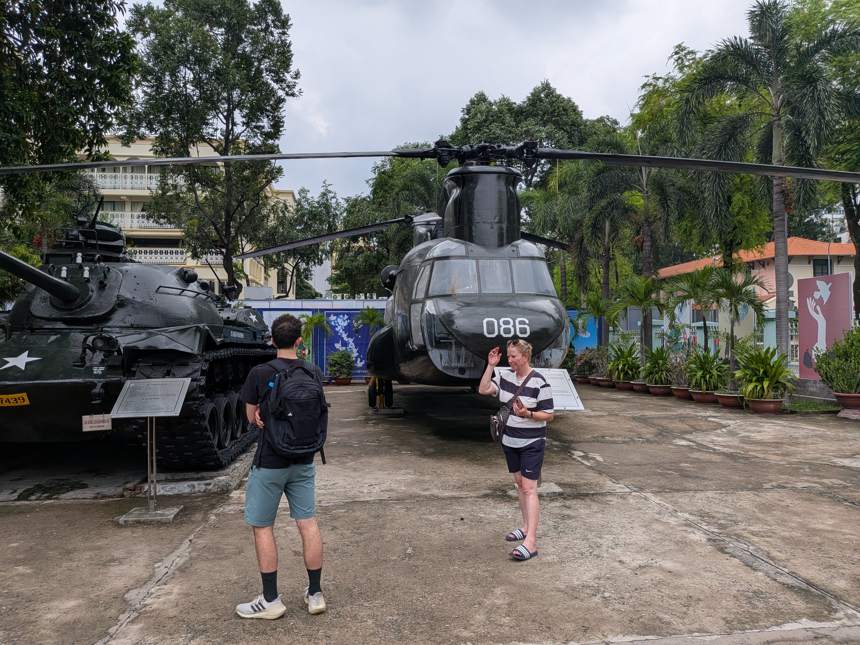 ベトナム戦争証跡博物館(ホーチミン市)の屋外展示エリアで、2人の旅行者が米軍のヘリコプター(番号086)と戦車の展示物を見学している様子。