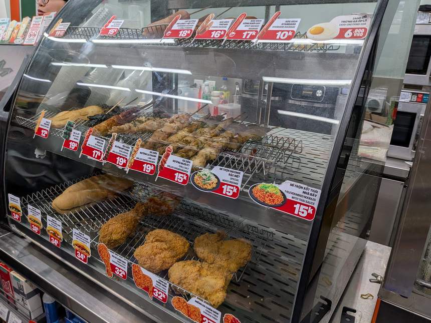 コンビニのホットフードケース。フライドチキン・串焼き・バインミーがドン建ての値札付きで並ぶ
