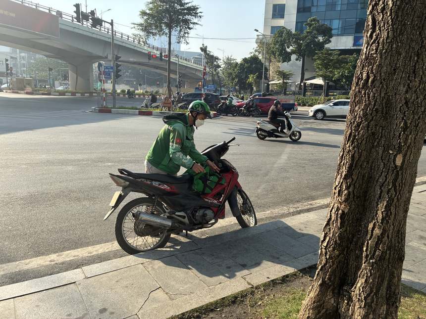 ベトナムの都市部の交差点付近で、配車サービスGrabの緑のジャケットを着たバイクドライバーがバイクのそばに立つ様子。会社員が週末などに行う副業の代表例。