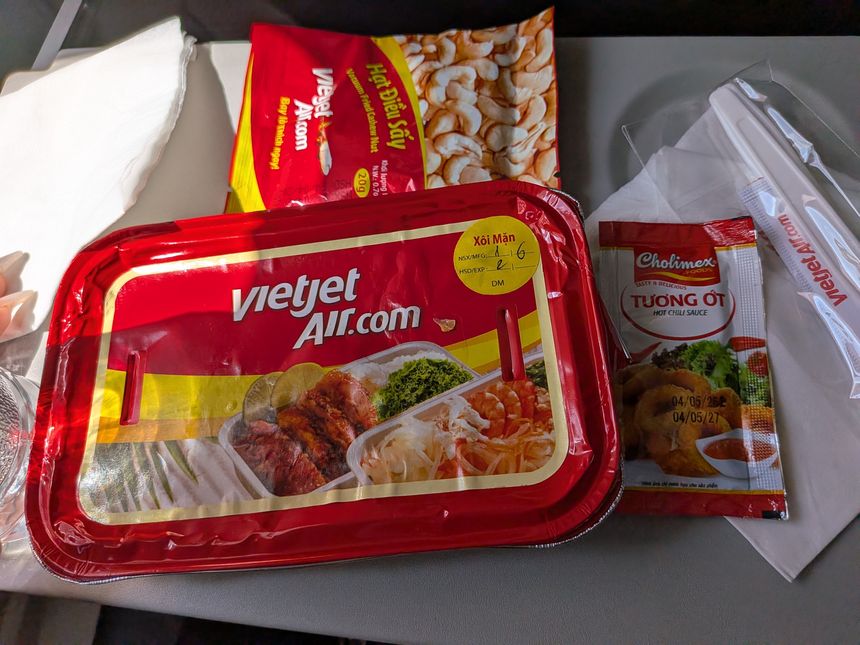 Vietjet Airの機内食トレイ。おこわ料理の弁当に、カシューナッツのスナックとCholimexのチリソースが添えられている。