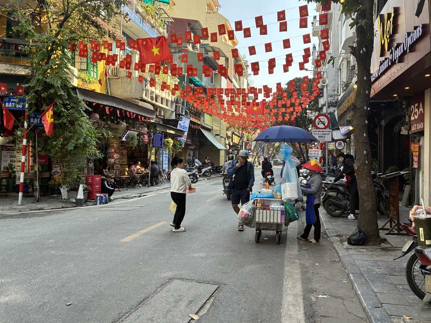 ベトナム国旗が連なるハノイ旧市街の通り。カートで物を売る行商人や歩行者が行き交い、フォーやカフェの店が並ぶ活気ある街並み。