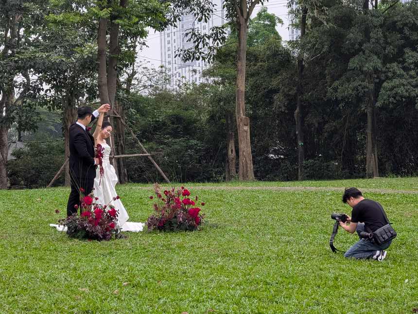 ベトナムの公園で、ウェディングドレスとタキシード姿のカップルが結婚式の前撮り撮影をしている。背景には高層ビルが見える。