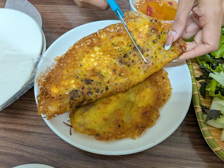 焼きたてのバインセオをハサミで切り分けているところ。左に袋入りのライスペーパー、右に生野菜のトレイ