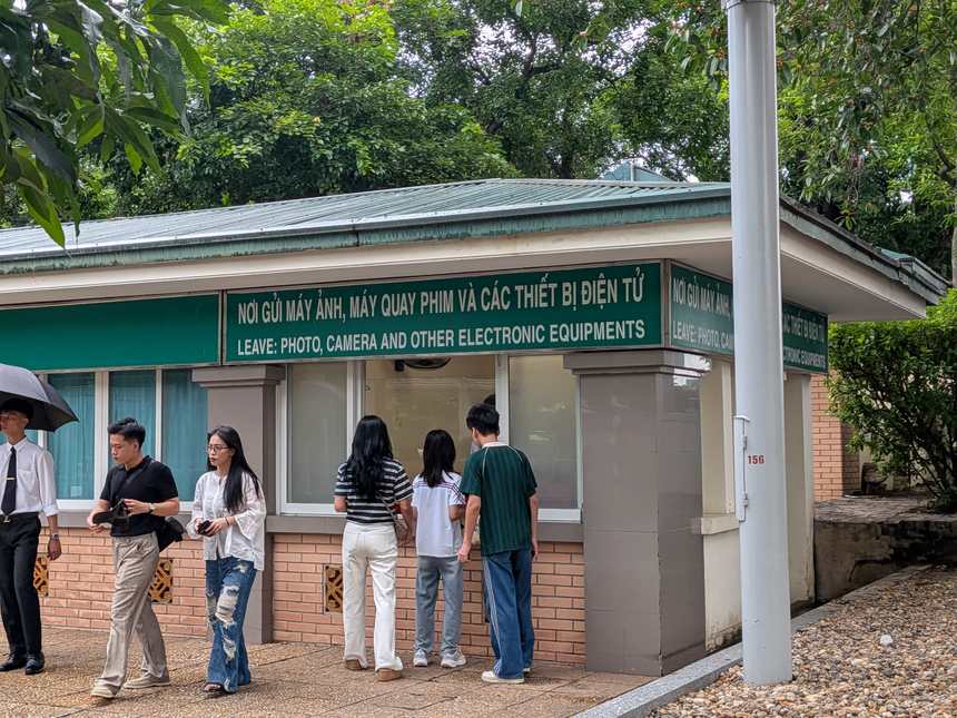 カメラや電子機器の預かり所。「NƠI GỬI MÁY ẢNH…／LEAVE: PHOTO, CAMERA AND OTHER ELECTRONIC EQUIPMENTS」と表示された窓口に見学者が並ぶ