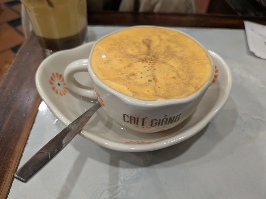 Cafe Giảngのホットのエッグコーヒー。白いカップに黄色い卵黄クリームがなみなみと乗り、スプーンが添えられている。