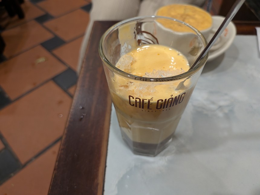 Cafe Giảngのアイスのエッグコーヒー。ロゴ入りのグラスに氷と卵クリームが入り、奥にホットのカップが見える。