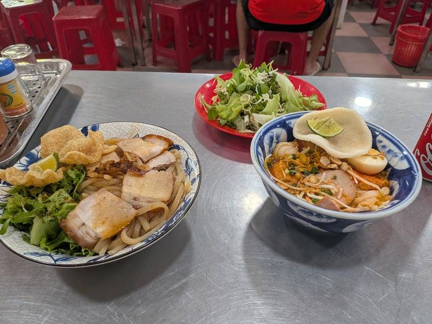 ミークアン1Aで並べたカオラウ（左）とミークアン（右）。カオラウは太い麺に豚肉と揚げた生地、ミークアンは黄色い麺にエビと卵、奥に生野菜の皿