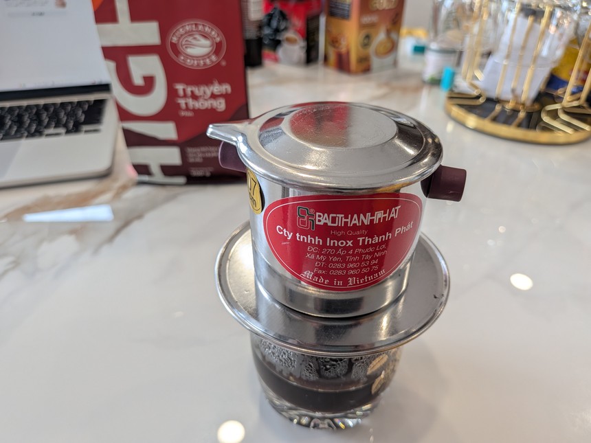 上ぶたをかぶせたフィンをグラスに載せたところ。本体にInox Thành Phátのラベル、後ろにハイランズコーヒーTruyền Thốngの袋