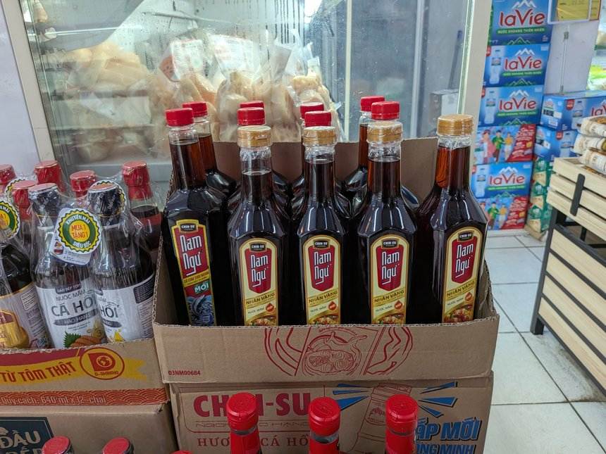 スーパーの売り場で段ボール箱に詰めて並べられた、赤いラベルのナムグー（Nam Ngư）のヌクマムの瓶