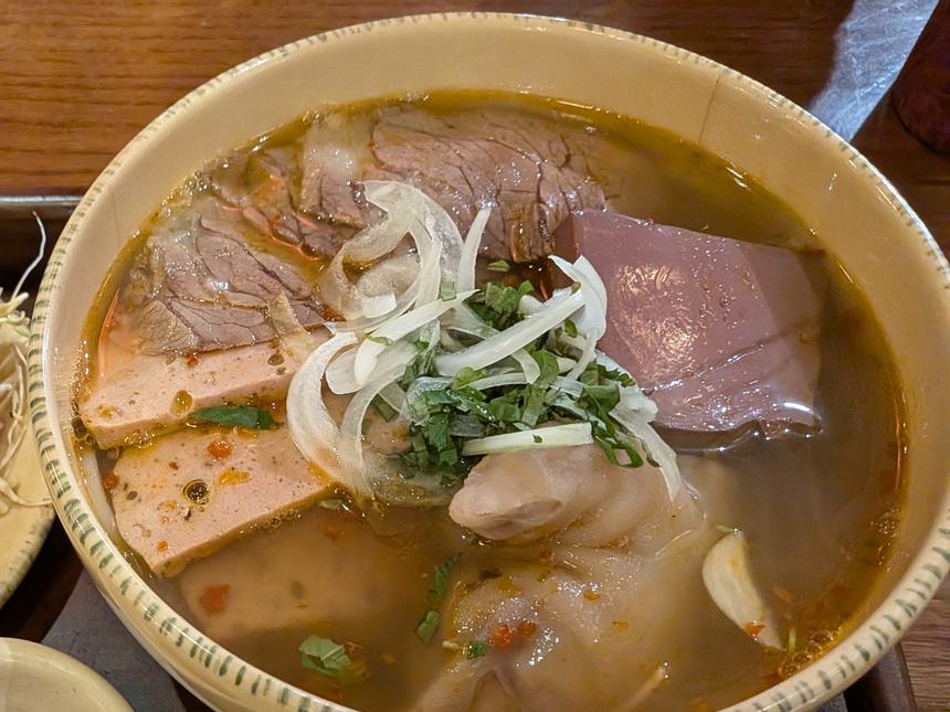ブンボーフエの丼。薄切りの牛肉、豚足、ピンク色のハムのような薄切り、赤紫色の四角い具、玉ねぎと刻んだ香草が、赤い油の浮いたスープに入っている