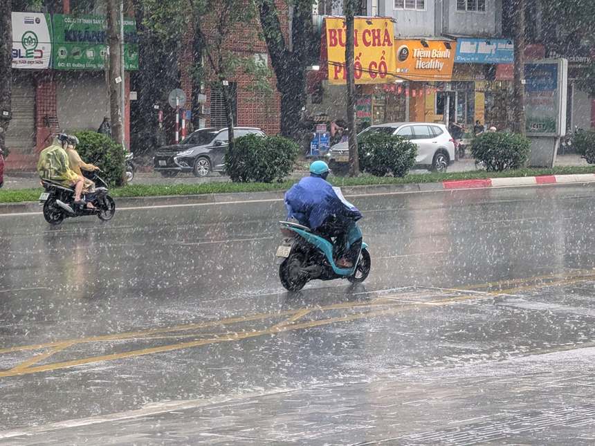 2026年7月19日昼のハノイの通り。激しい雨で路面に雨しぶきが立ち、レインコートの人がバイクで走る