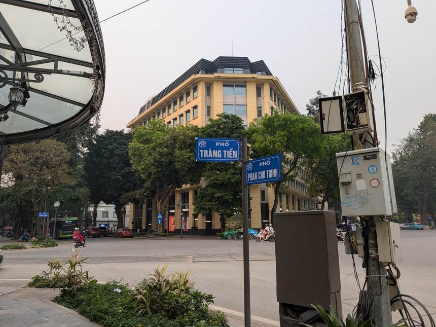 「PHỐ TRÀNG TIỀN」と「PHỐ PHAN CHU TRINH」の青い通りの名前の標識。後ろに黄色い外壁の建物と街路樹