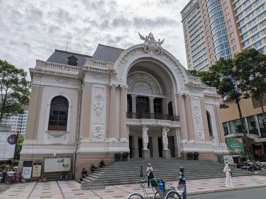 日中のホーチミン歌劇場の正面。アーチの上に羽のある像、入口の両わきに女性の像が立ち、右に高いビルが見える