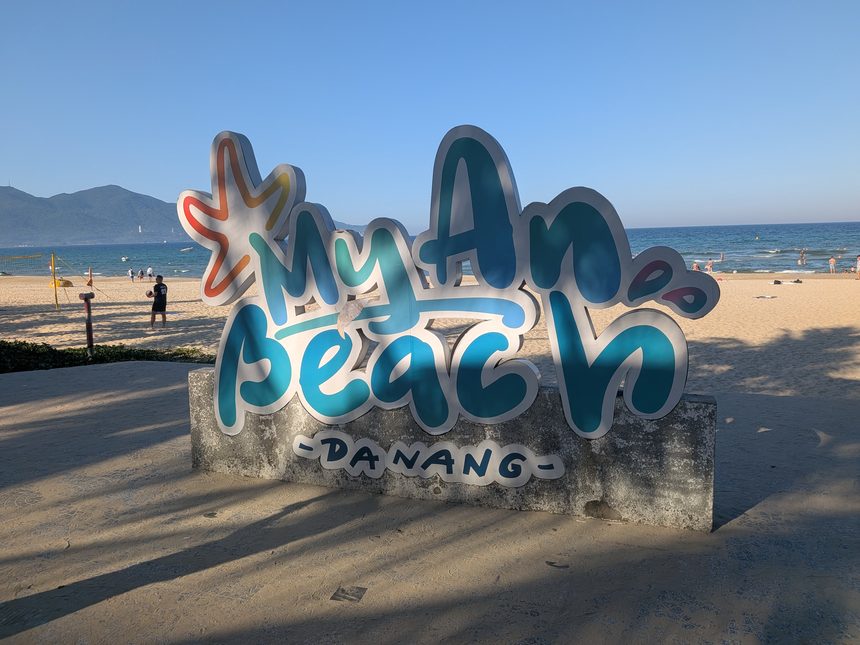 浜の手前に立つ「My An Beach – DANANG」の看板。後ろに砂浜と海、左の奥に山が見える