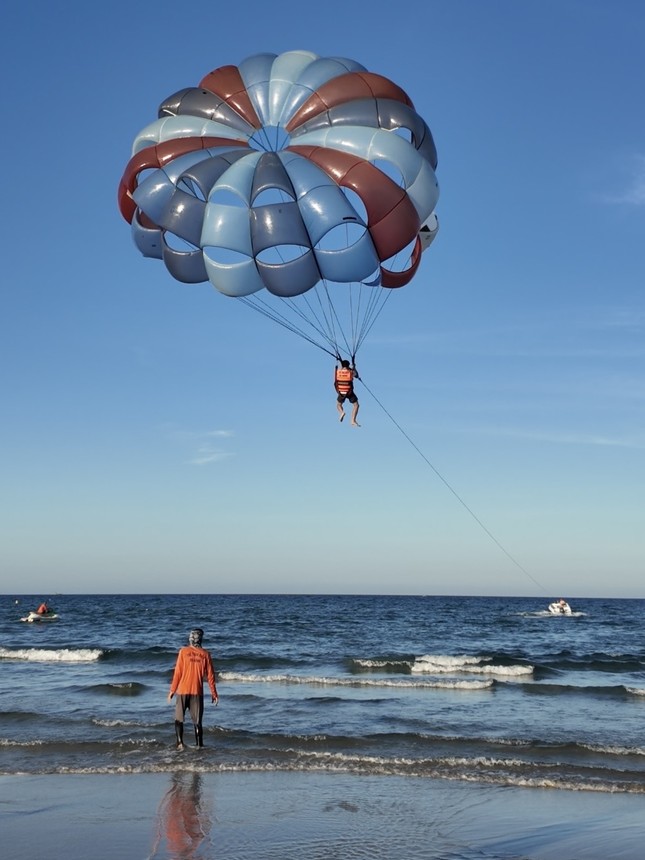 青空に浮かぶパラセーリング。ロープの先の沖にボートがあり、波打ち際にオレンジの服の人が立っている